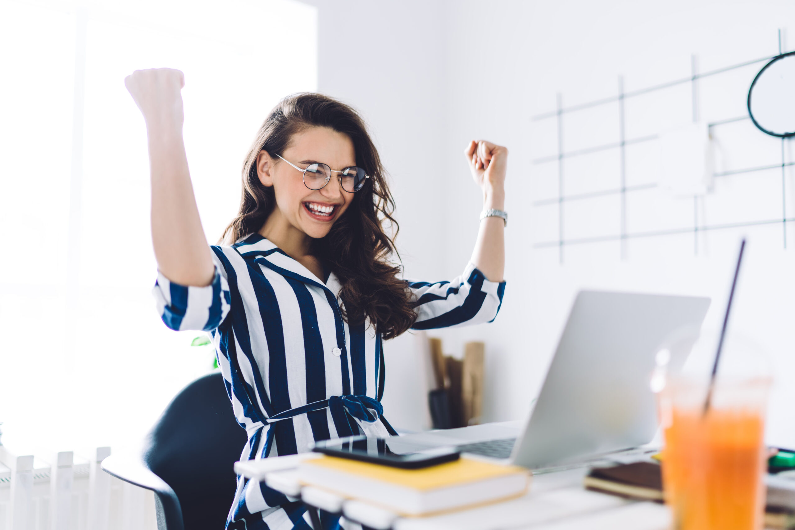 A happy woman with glasses celebrating success while working on her laptop in a bright office.
